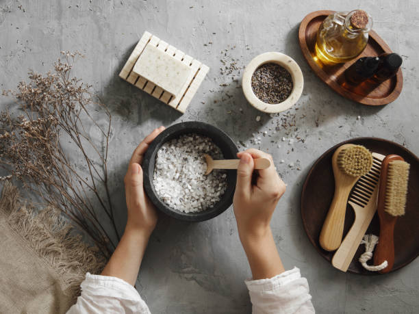 showing two hands of a woman mixing bath salts with lavender in a dish with other smaller dishes around with tools and ingredients to assist with this craft